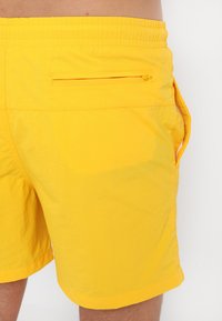 Shorts de bain jaunes en tissu léger. Les caractéristiques incluent une taille élastique et une poche arrière zippée. Texture lisse.