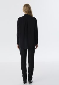 Schwarzes Langarmshirt mit hohem Kragen, asymmetrischem Saum und Rückenschlitz, kombiniert mit schwarzen taillierten Hosen und High Heels.