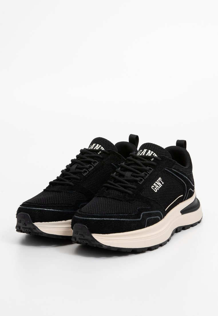 Baskets de sport noires avec une tige en mesh et en daim, équipées de lacets noirs, d'un logo blanc et d'une semelle intermédiaire épaisse beige avec une semelle extérieure texturée.