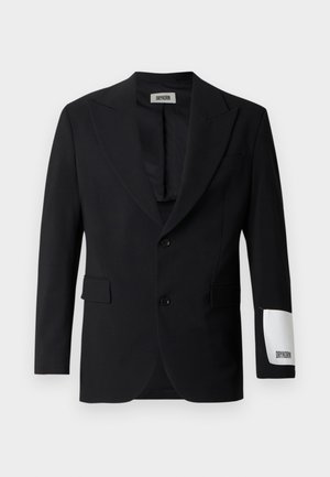 Zwart colbert van gladde stof, voorzien van een aangeknipte reverskraag, twee knopen en zijzakken. Opvallend wit label op de mouw met de tekst "DRYKORN."