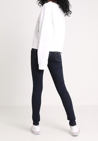 Bílá zkrácená mikina s volnými rameny, v kombinaci s tmavě modrými skinny džíny a bílými plátěnými teniskami. Hladká textura a minimalistický design.