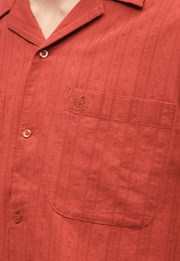 Κοντινό πλάνο ενός ατόμου που φοράει πουκάμισο με κουμπιά σε σκουριαστό χρώμα, με υφή και τσέπη στο στήθος που φέρει κεντημένο λογότυπο.