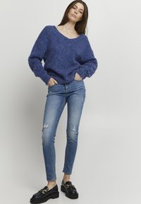 Pull en tricot bleu avec un décolleté large et une coupe ample, associé à un jean skinny bleu clair avec des détails déchirés, et des mocassins noirs.