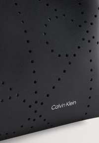 Μαύρη δερμάτινη επιφάνεια με ομοιόμορφα διατεταγμένες οπές που σχηματίζουν ένα διακριτικό μοτίβο, με το λογότυπο "Calvin Klein" σε λευκό κείμενο.