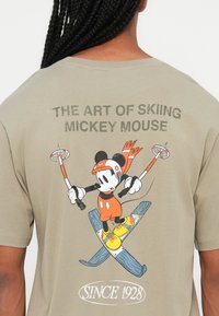 Camiseta verde oliva con un gráfico de dibujos animados de Mickey Mouse esquiando, con acentos rojos, bastones de esquí y el texto "EL ARTE DEL ESQUÍ DESDE 1928."