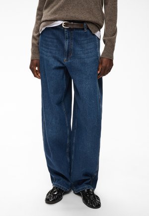 Person trägt lockere dunkelblaue Jeans, braunen Pullover, weißes Hemd eingesteckt, schwarzen Gürtel und schwarze Lederschuhe und steht vor weißem Hintergrund.