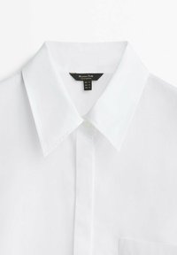 Camicia bianca con colletto classico, chiusura a bottoni e una tasca sul petto. Realizzata in tessuto liscio, con etichetta nera contenente le istruzioni per la cura.