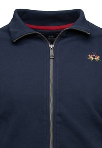 Námořnická zipová bunda vyrobená z měkké tkaniny s vysokým límcem, stříbrným zipem a červenou podšívkou, s vyšitým logem dvou koní.