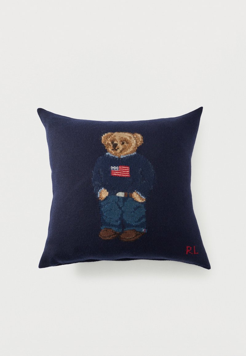 Μαξιλάρι ναυτικού μπλε με πλεκτή αρκουδάκι που φοράει τζιν, καφέ παπούτσια και πουλόβερ με αμερικάνικη σημαία, με τα αρχικά "RL" σε κόκκινο.