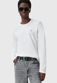 Weißes Langarmhemd mit kleinem schwarzem Logo, kombiniert mit grauen Jeans und einem schwarzen Gürtel mit Metallösen und einer silbernen Schnalle.