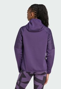 En kvinna med flätat hår som bär en lila hoodie och mönstrade leggings ses bakifrån, vilket framhäver designen och passformen på hoodien.