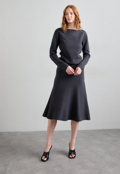 Dunkelgraues Strickkleid mit langen Ärmeln und volanartigem Saum, kombiniert mit schwarzen Open-Toe-Schuhen. Glatte Textur und minimalistisches Design.