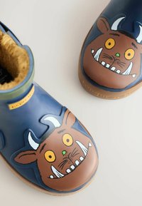 Blaue Gummistiefel mit einem braunen Monster-Gesichtdesign, das große Augen, weiße Zähne und ein texturiertes Plüschfutter aufweist. Braune Gummisohlen.