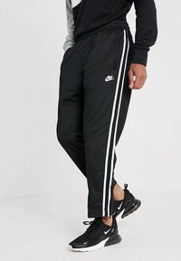 Personen bär svarta Nike träningsbyxor med vita sidostrecker och svarta Nike sneakers med vita sulor och logotyper.