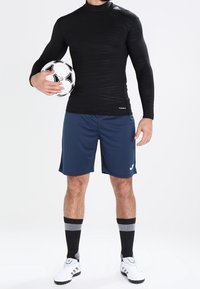 Czarna długorękawowa sportowa bluzka z teksturowanym wzorem, w zestawie z granatowymi szortami. Model trzyma czarno-białą piłkę nożną, ma na sobie czarne, sięgające kolan skarpetki.