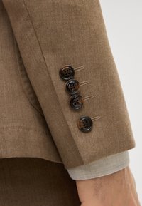 Brauner maßgeschneiderter Blazerärmel mit vier dunkel gefärbten Knöpfen, die ein kreisförmiges Design aufweisen, genäht mit kontrastierendem Faden und einem leichten Stoffunterlayer.