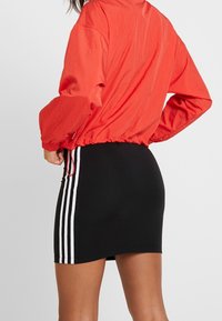 Röd lättviktsjacka med elastisk nederkant över svart åtsittande kjol med vita sidostråkar, slät textur och avslappnad design.