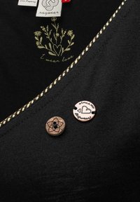 Svart tygnära med två knappar, blommönster, hjärtillustration och text som lyder "Jag bär kärlek" och "sedan 1997 ragwear."