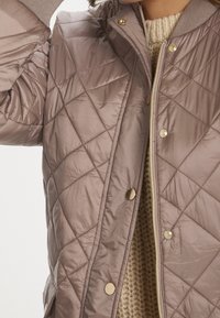 Prošívaná taupe bunda s kovovými detaily, vybavená zipem a patentkami. Texturovaná vnitřní vrstva se ukazuje z rozepnutého předního dílu.