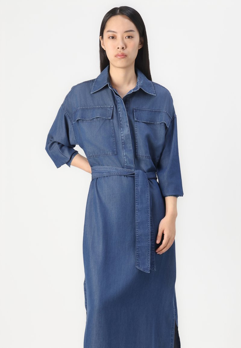 Robe chemise en denim avec une taille cintrée, des manches longues, des poches poitrine et une silhouette droite. Couleur bleu clair avec une texture lisse.