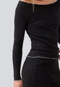 Schwarzes, geripptes Longsleeve-Oberteil mit Off-Shoulder-Design. Verfügt über weiße Rüschendetails am Kragen und Saum, die eine strukturierte Note verleihen.