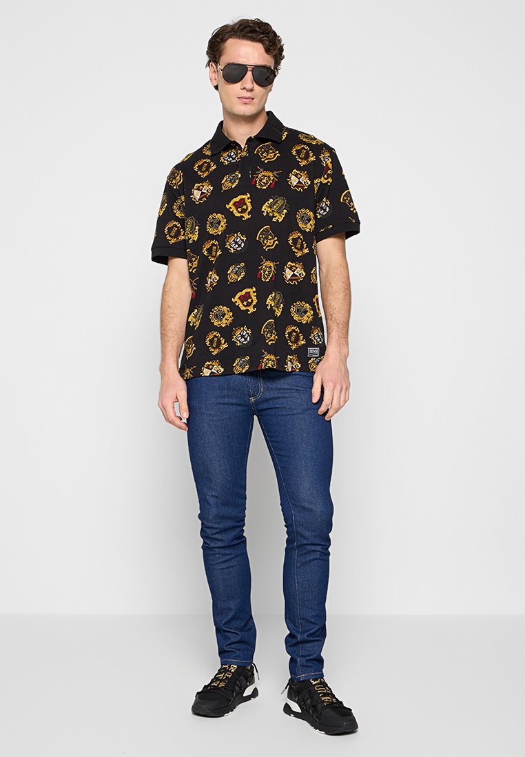 Schwarzes Polohemd mit goldenen und roten Mustern, kombiniert mit blauen Jeans. Das Modell trägt Sonnenbrillen und schwarze Schuhe mit gelben Akzenten.