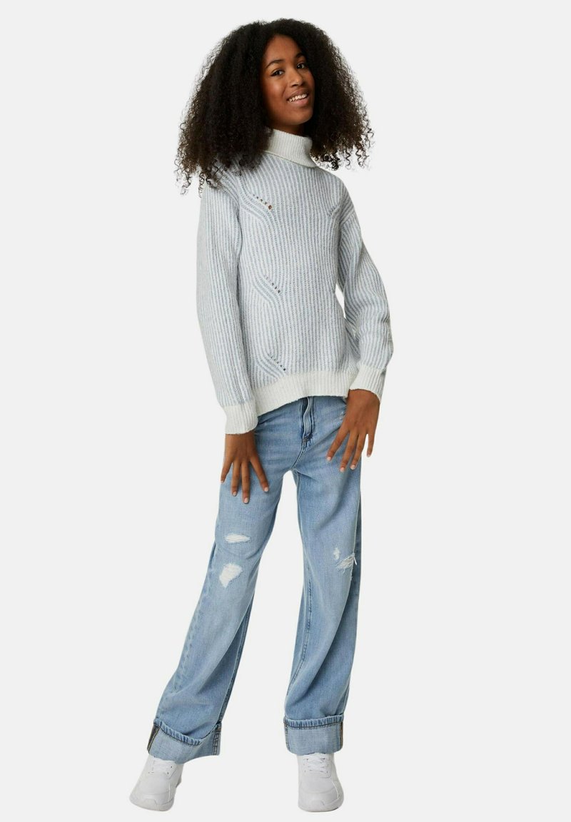 Pull en maille côtelée gris clair avec col montant, associé à un jean déchiré bleu clair et des baskets blanches. Le modèle pose les mains sur les hanches.