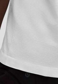 Weißes Sportshirt mit horizontaler Rippstruktur und abgerundetem Saum, kombiniert mit schwarzen Leggings, die am Bund ein Logo tragen.