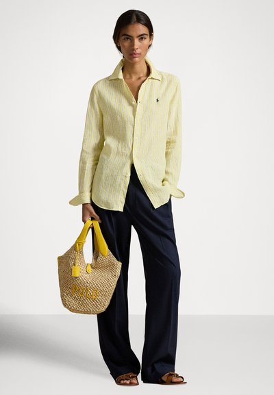 Sieviete, tērpusies dzeltenā svītrainā kreklā ar pogām un brīvās piegriezuma tumši zilās biksēs, rokā turot austu dzeltenas roktura somu un valkājot brūnas sandales.