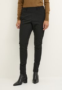 Pantalons noirs ajustés avec une texture lisse, dotés d'une taille haute et de détails subtils le long des coutures, associés à des bottines noires à talons.