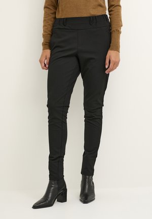 Pantalons noirs ajustés avec une texture lisse, dotés d'une taille haute et de détails subtils le long des coutures, associés à des bottines noires à talons.