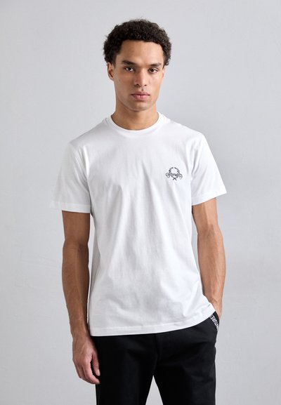 Camiseta blanca de algodón con mangas cortas, cuello redondo y un pequeño gráfico impreso en el lado izquierdo del pecho. Acentos negros en el dobladillo de la camiseta.