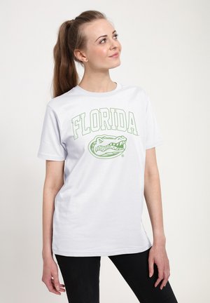 Ung kvinde med brunt hår i hestehår, iført hvid t-shirt med grøn Florida-tekst og alligatorlogo, ser opad mod en ensartet baggrund.