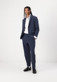 Marineblauw pak met een colbert met één knoop, bijpassende broek en een wit shirt. Glanzende zwarte instappers maken het geheel af.