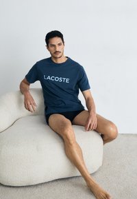 Tamno plava pamučna majica s logom "LACOSTE" u svijetlo plavoj boji, nosi se s crnim šortsevima, sjedi na krem boji, skulpturalnoj stolici.