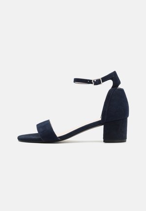 Sandale en suède bleu marine avec une large lanière avant, une lanière de cheville réglable et un talon carré. Texture lisse avec un design minimaliste.