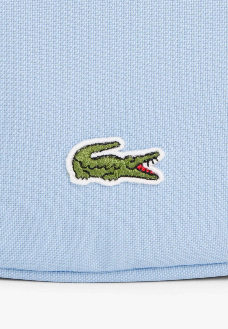 Logo de crocodile vert brodé avec une gueule rouge ouverte sur un fond de tissu bleu clair texturé.