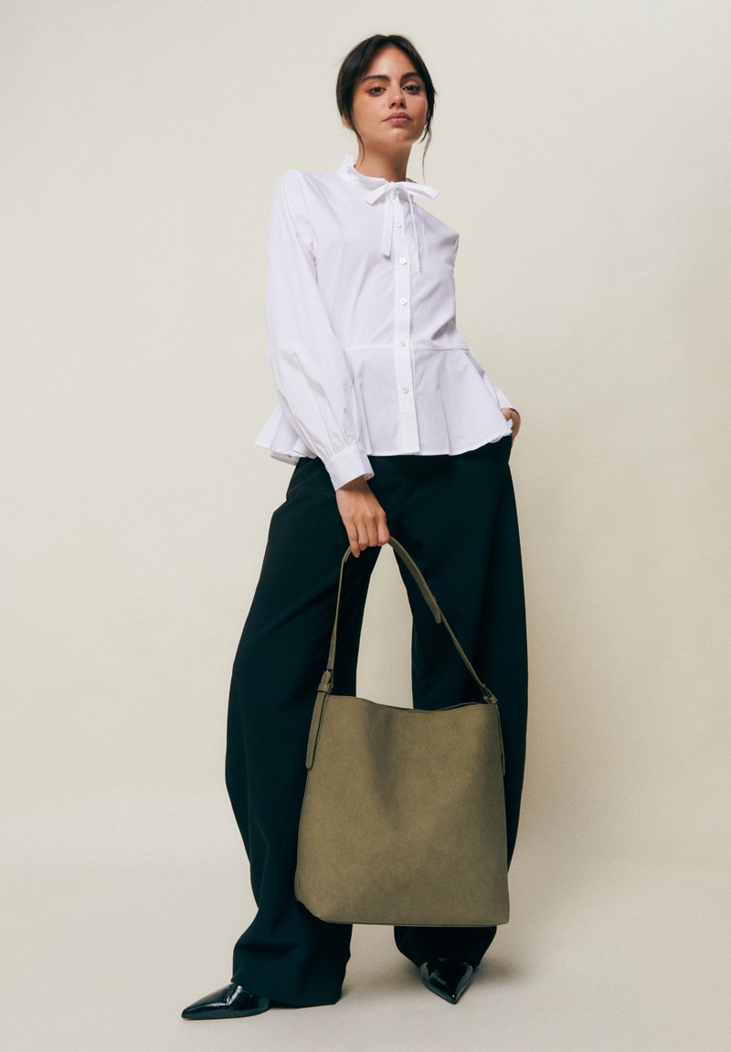 Mujer con una blusa blanca abotonada y pantalones anchos negros, sosteniendo un gran bolso verde oliva por la correa, sobre un fondo liso.