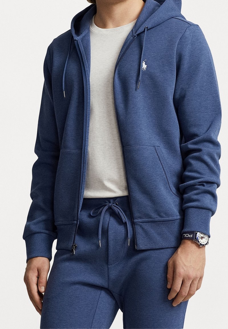 Homme portant un sweat à capuche bleu zippé avec un logo blanc brodé, un pantalon de survêtement bleu assorti, un t-shirt blanc et une montre avec un bracelet foncé.