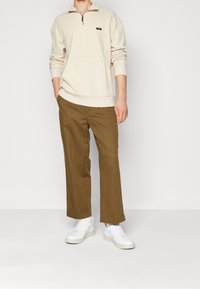 Sudadera beige con cremallera y bolsillo delantero, combinada con pantalones amplios marrones y zapatillas blancas. Textura de tela suave y ajuste relajado.