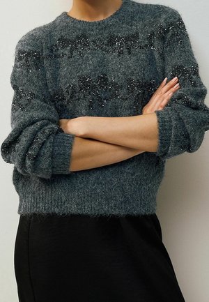 Personne portant un pull gris foncé duveteux avec des motifs en sequins noirs, bras croisés, associé à une jupe noire sur un fond uni.