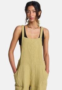 Φόρμα σε ανοιχτό πράσινο ελιάς, κατασκευασμένη από υφή υφάσματος με φαρδιές τιράντες, τσέπες μπροστά και χαλαρή γραμμή, σε συνδυασμό με μαύρο τοπ από κάτω.