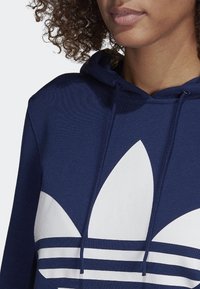 Tummansininen huppari, jossa on valkoinen kolmi-nauha design ja logo edessä. Hupparissa on naruhuppu ja teksturoitu kangas.