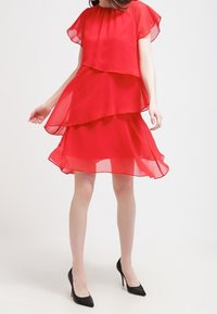 Robe en chiffon rouge avec un design superposé, des manches évasées courtes et un col rond. Associée à des chaussures à talons hauts noires.