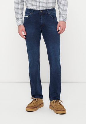 Mann trägt dunkelblaue Jeans, hellbraune Wildlederschnürschuhe und ein kariertes Langarmhemd und steht vor einem einfarbigen weißen Hintergrund.