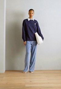 Ναυτική μπλούζα με λευκό γιακά, ανοιχτό μπλε παντελόνι σε φαρδιά γραμμή και λευκή τσάντα tote. Το μοντέλο στέκεται σε ξύλινο δάπεδο μπροστά από ουδέτερο τοίχο.