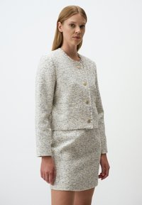 Giacca in tweed leggermente strutturata, crema e blu, con taglio corto, scollatura rotonda e bottoni dorati. Abbinata a una gonna coordinata.