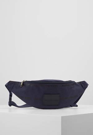 Sac banane Calvin Klein Jeans bleu marine avec fermeture éclair et sangle ajustable, posé sur une surface blanche.