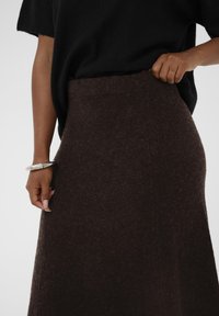 Rjava volnena krila z oprijetim pasom, gladko teksturo in razširjenim dizajnom. Dopolnjena z ohlapnim črnim puloverjem in srebrnim zapestnico.