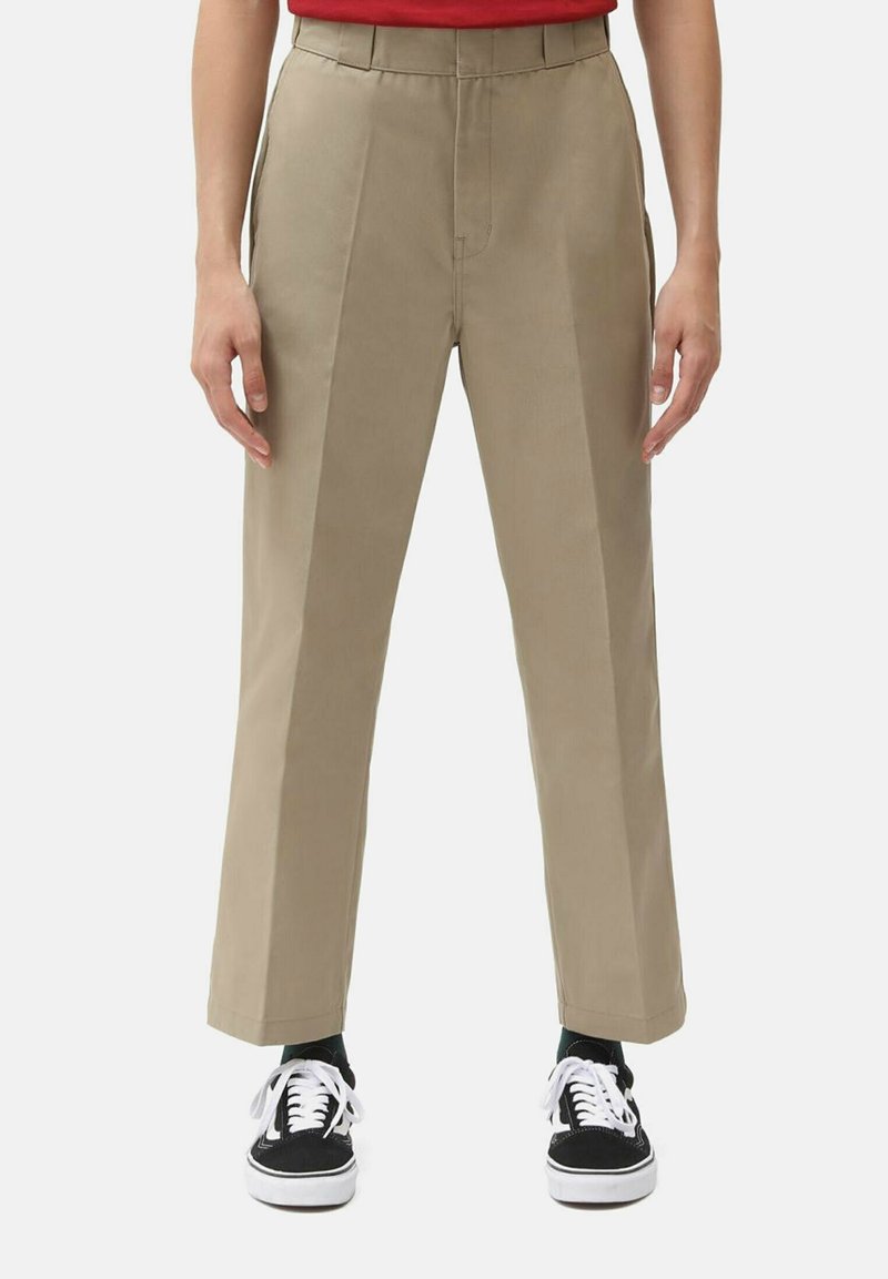 Beige high-waisted pants with a straight leg design, featuring pleats and side pockets, paired with black sneakers and white laces.